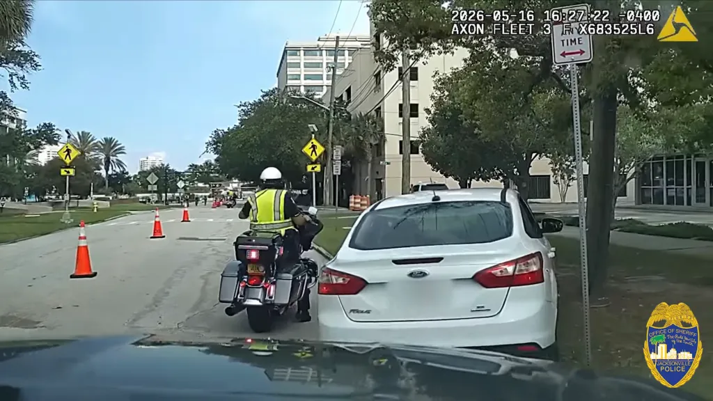 Dashboard camera footage showing a police motorcyclist and a white car on the right, with traffic cones in the road.