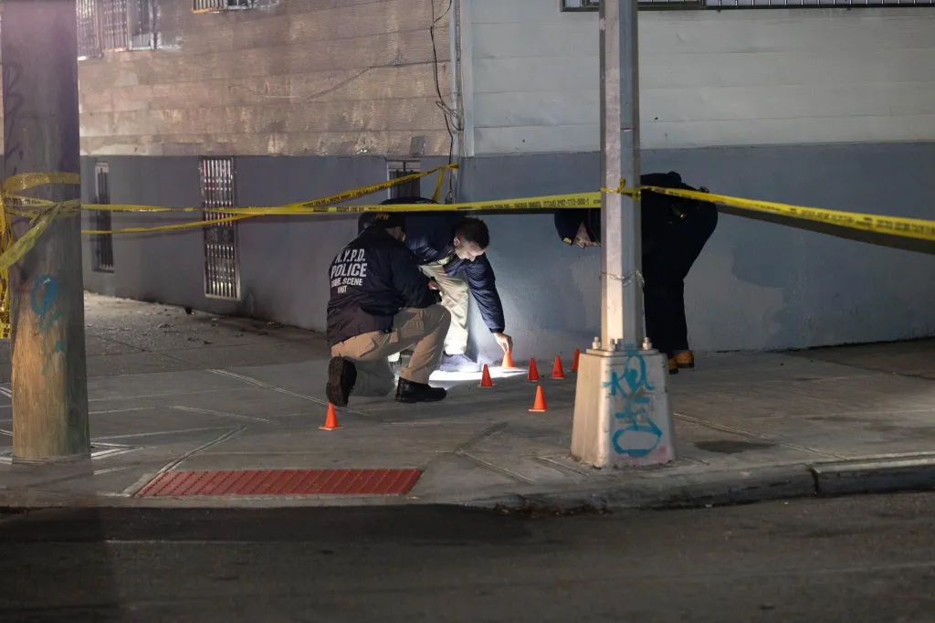 Police Crime Scene Unit officers investigate a multiple shooting scene, with several orange cones marking evidence on the sidewalk.