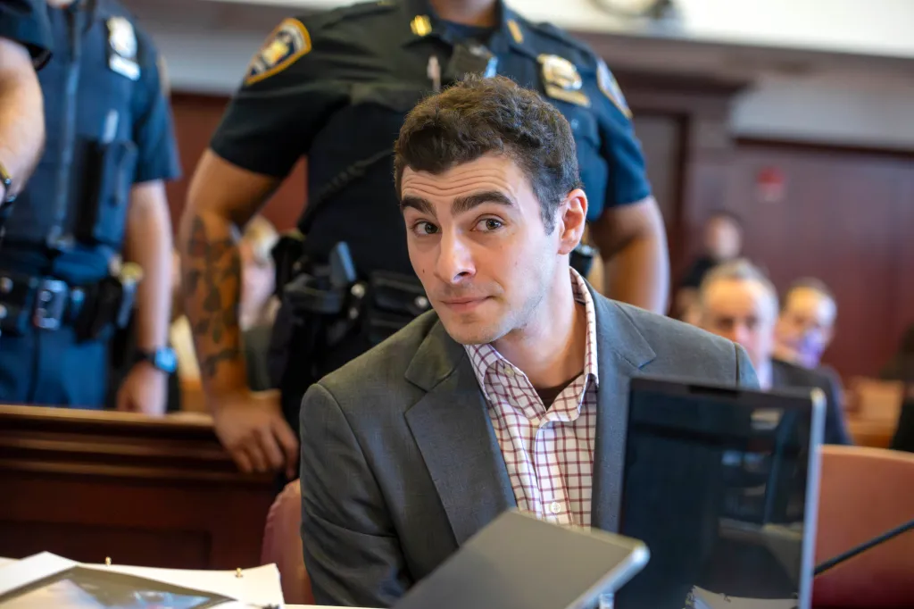 Luigi Mangione, an Ivy League graduate, looks toward the camera while wearing a gray suit and plaid shirt in a courtroom.