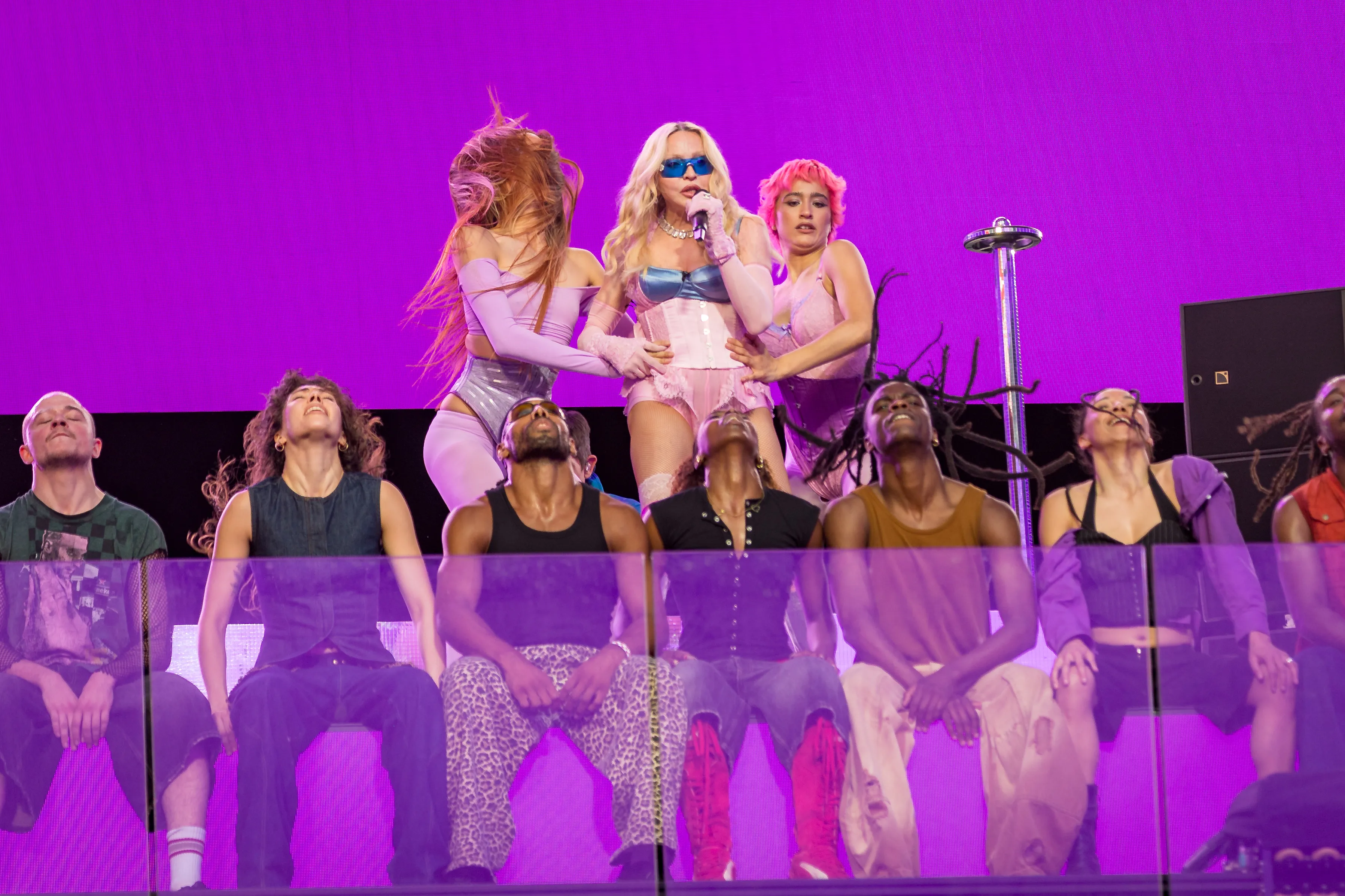 Popstar Madonna performs a pop-up concert, with two backup dancers next to her and others sitting below, against a bright purple background.