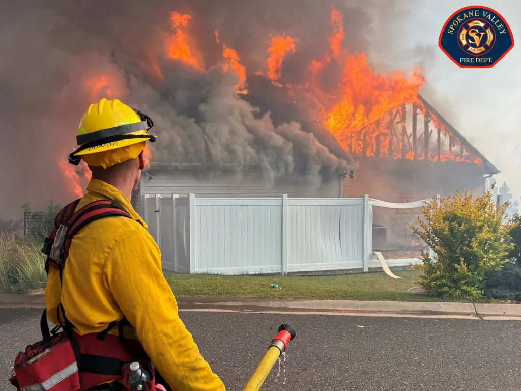 A firefighter in yellow gear watching a house engulfed in flames and smoke.