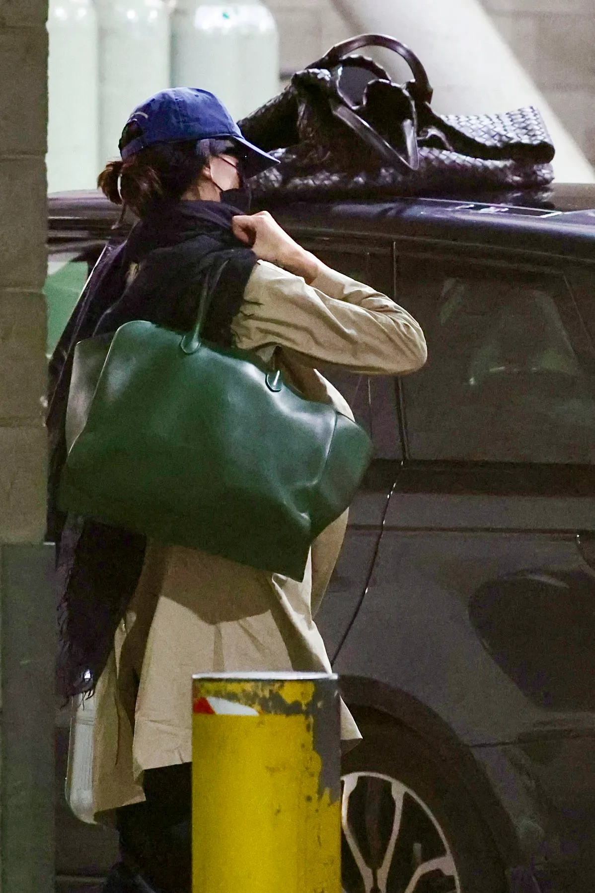 Kendall Jenner in a beige coat, black scarf, blue cap, black facemask, and carrying a large green bag.