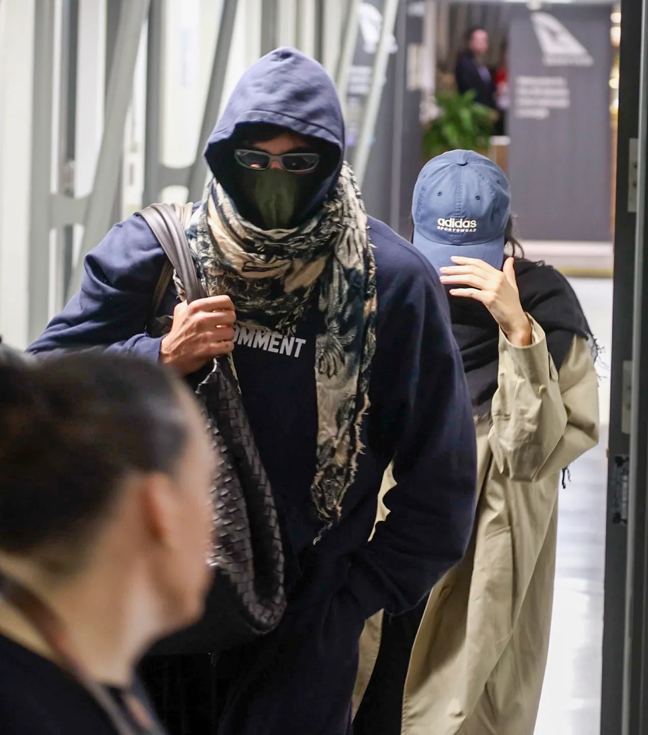 Jacob Elordi in a dark blue hoodie, beige scarf, black facemask, and sunglasses, with Kendall Jenner in a beige coat and blue cap, departing Brisbane Airport.