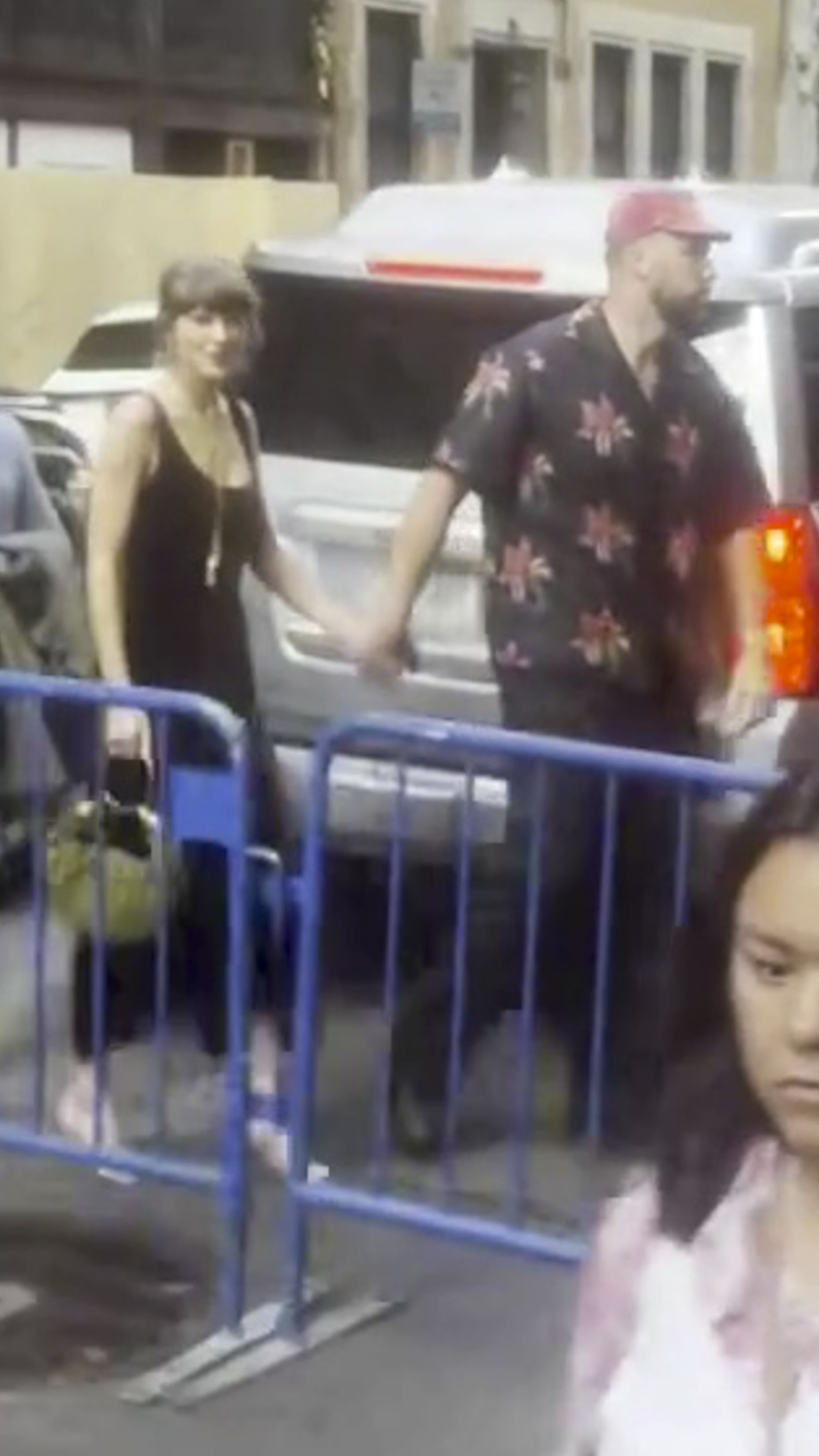 Taylor Swift and Travis Kelce holding hands while walking, with a blue barrier in front of them.