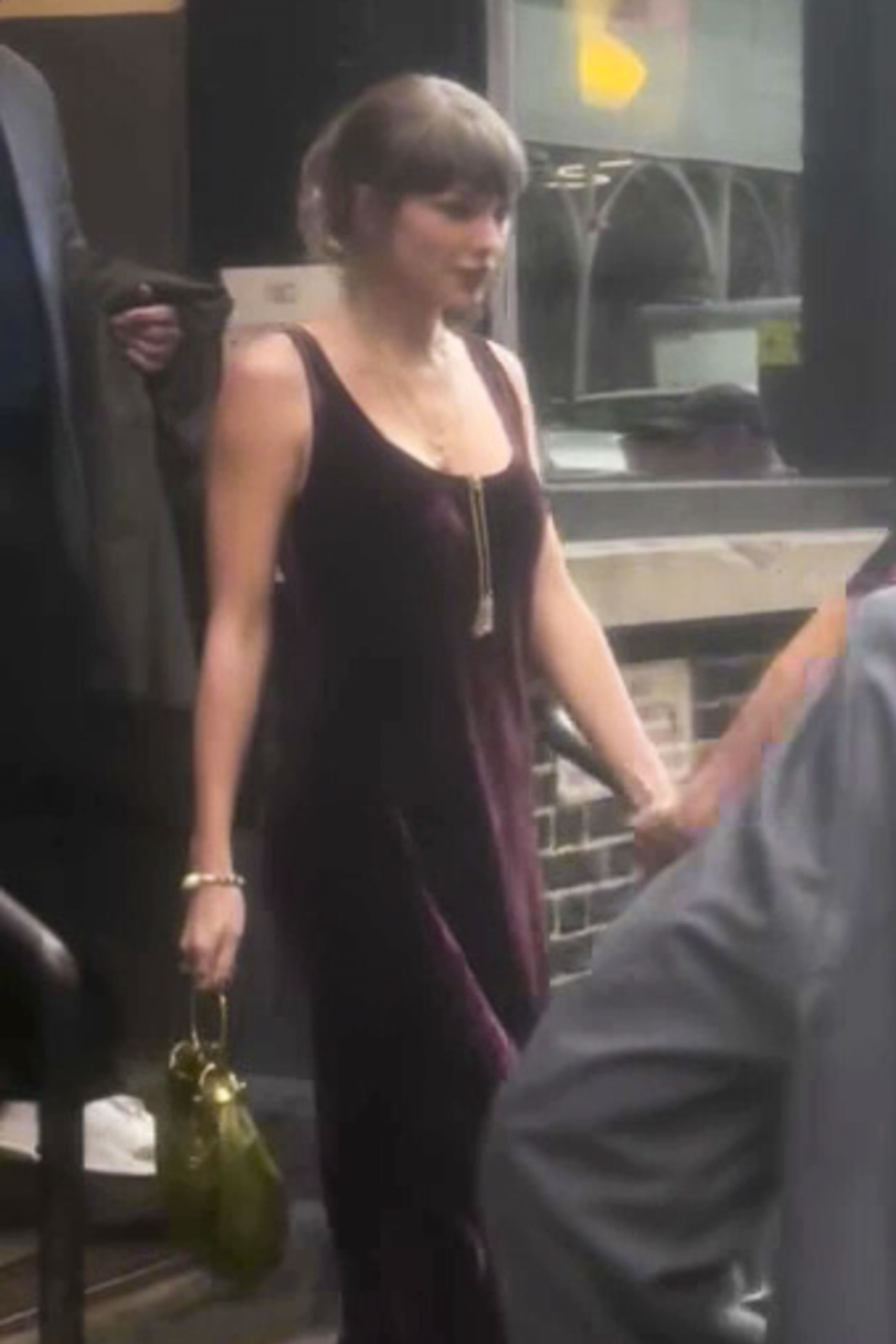 Taylor Swift in a dark-colored sleeveless dress with a long necklace and a green purse.