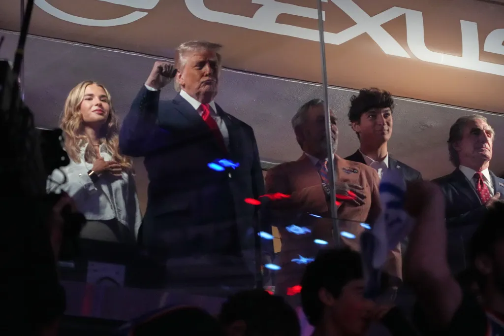 Donald Trump and others in a box suite stand for the National Anthem.