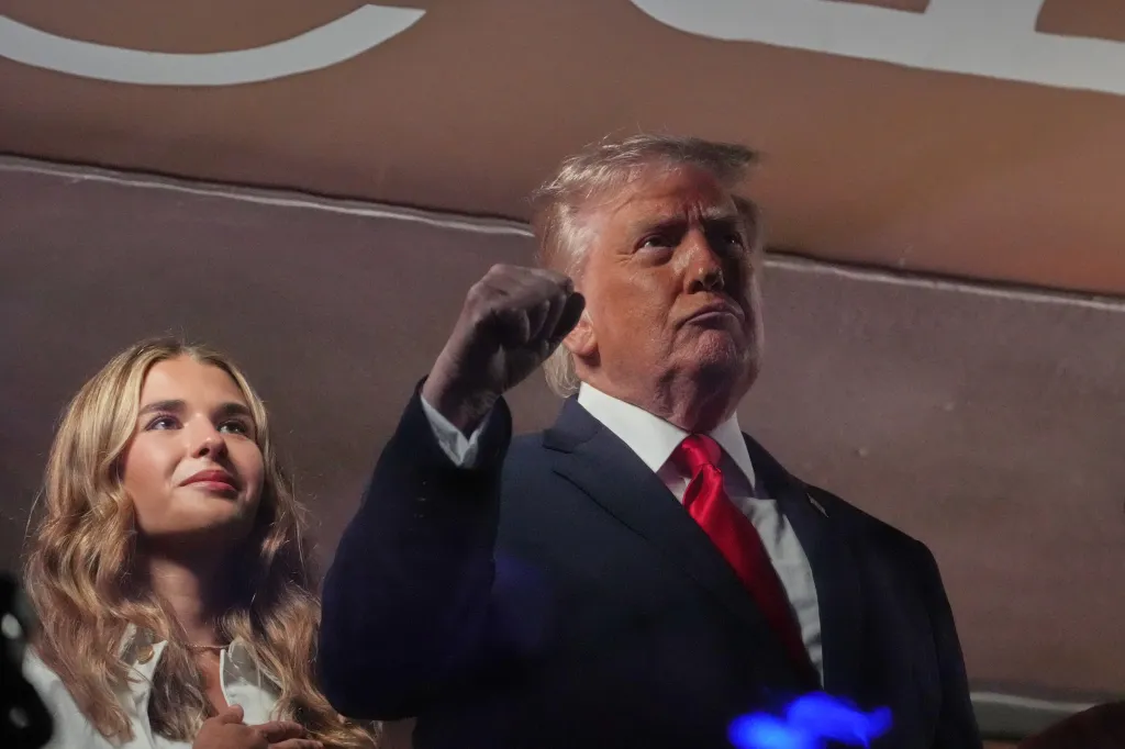 Donald Trump, wearing a suit and red tie, with his fist raised next to Kai Trump, who has her hand over her heart.