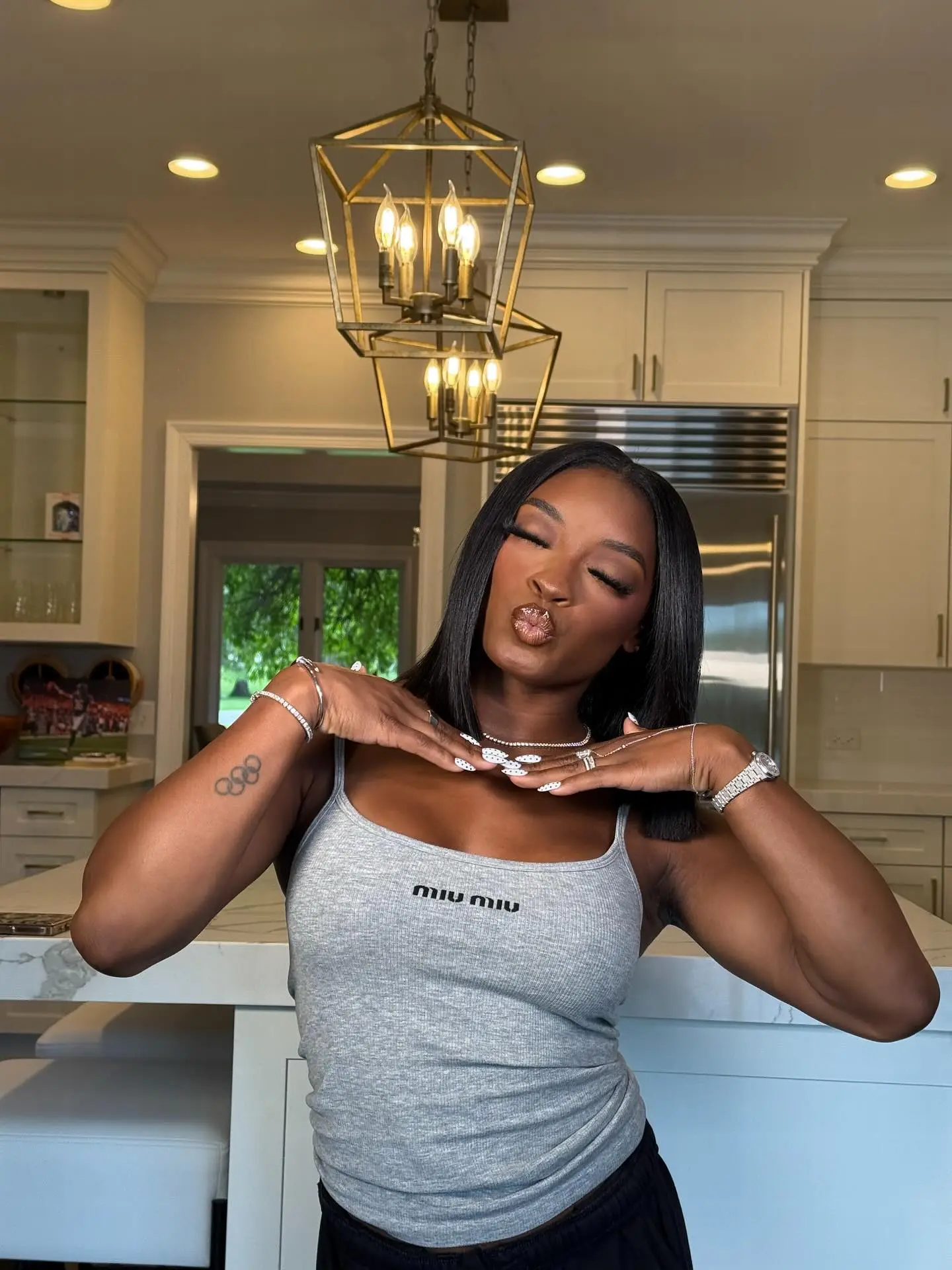 Simone Biles making a kissing face while wearing a gray Miu Miu tank top and showing her Olympic rings tattoo.