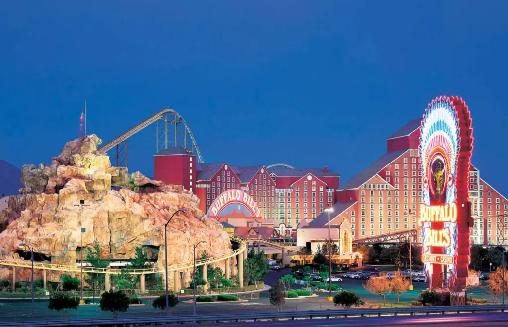 Buffalo Bill's Resort and Casino illuminated at dusk, with a roller coaster winding around a rock formation and extending towards the red-sided hotel.