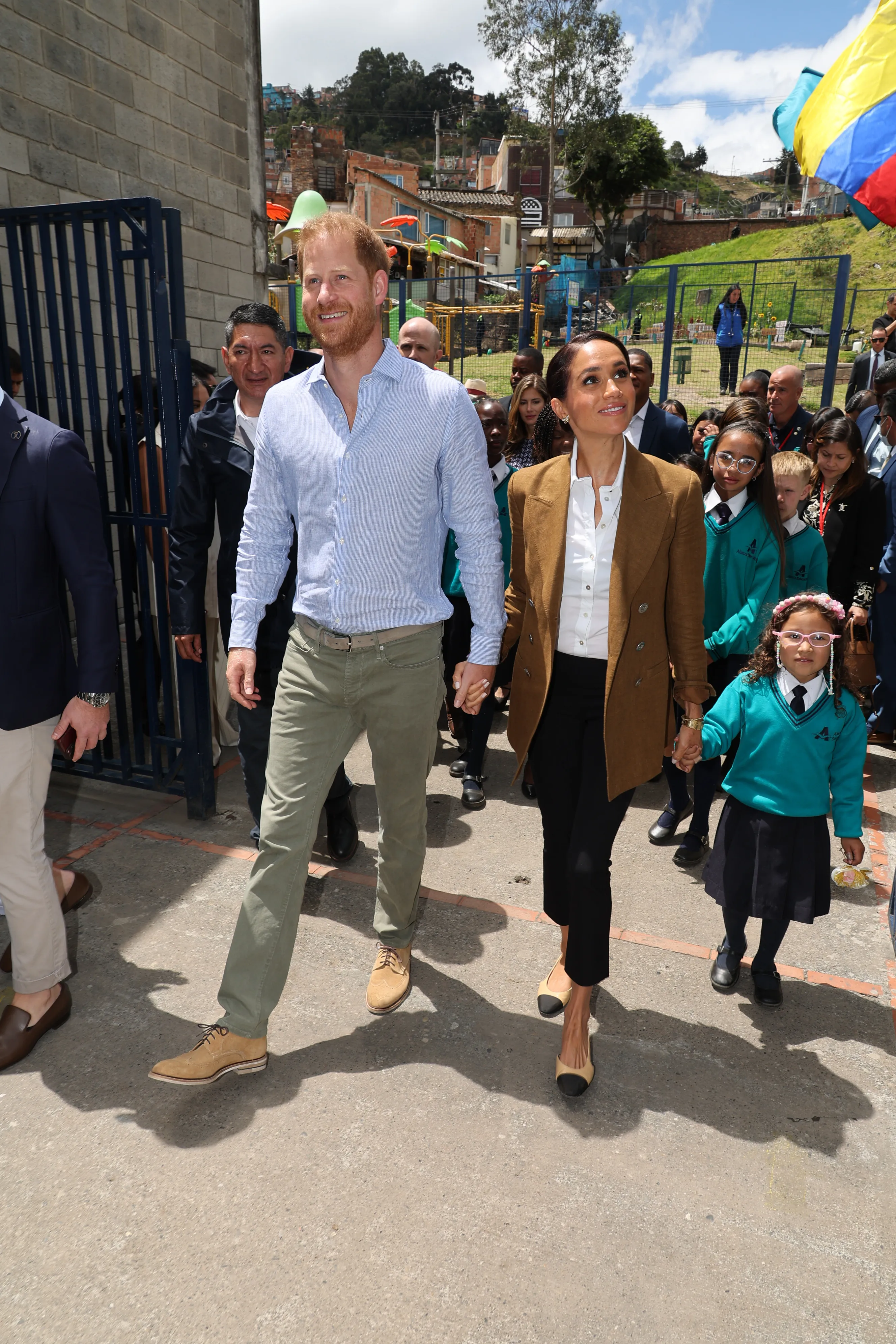 Prince Harry and Meghan Markle holding hands while walking through a school with students.