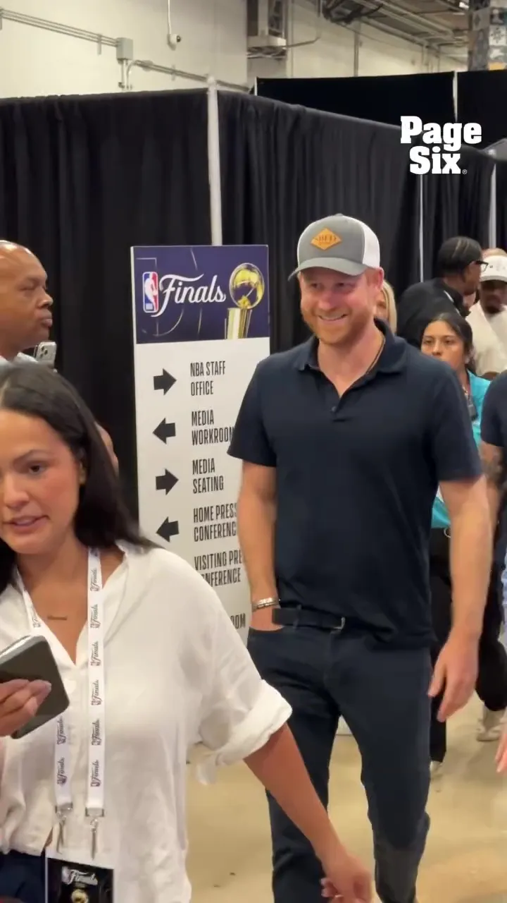 Prince Harry smiling in a blue polo shirt and a gray and white cap, walking past a sign for NBA Finals staff offices.