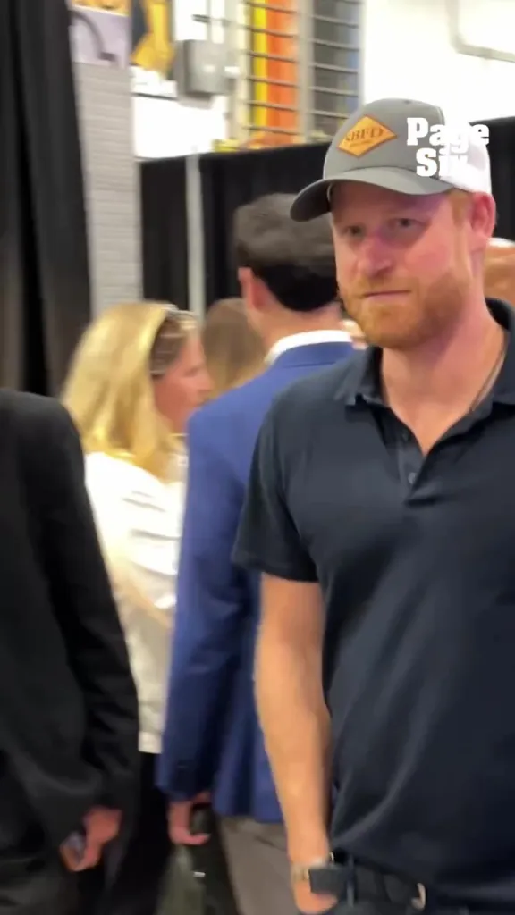 Prince Harry wearing a gray hat with a red and gold logo and a navy blue polo shirt.