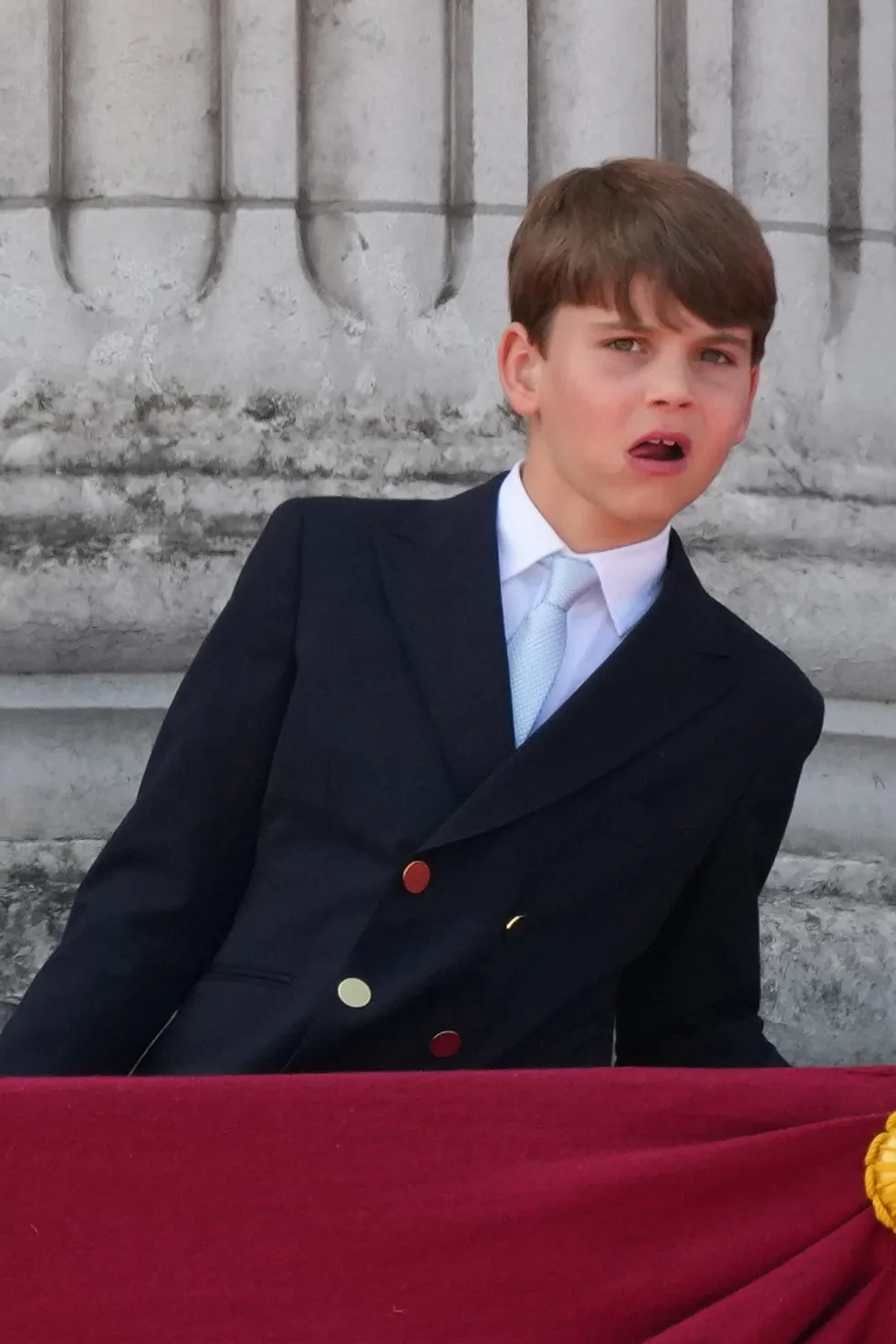 Prince Louis leans over a red velvet railing with his mouth slightly open, looking at the crowd from the balcony of Buckingham Palace.