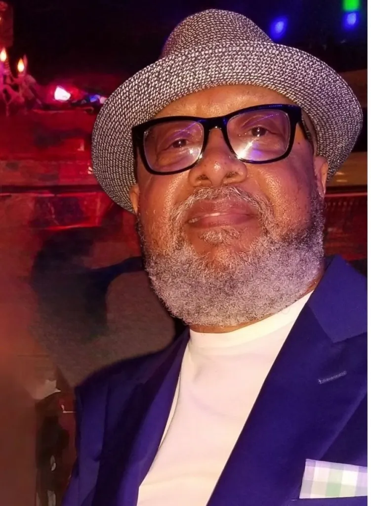 A man with a salt and pepper beard and fedora smiles at the camera.