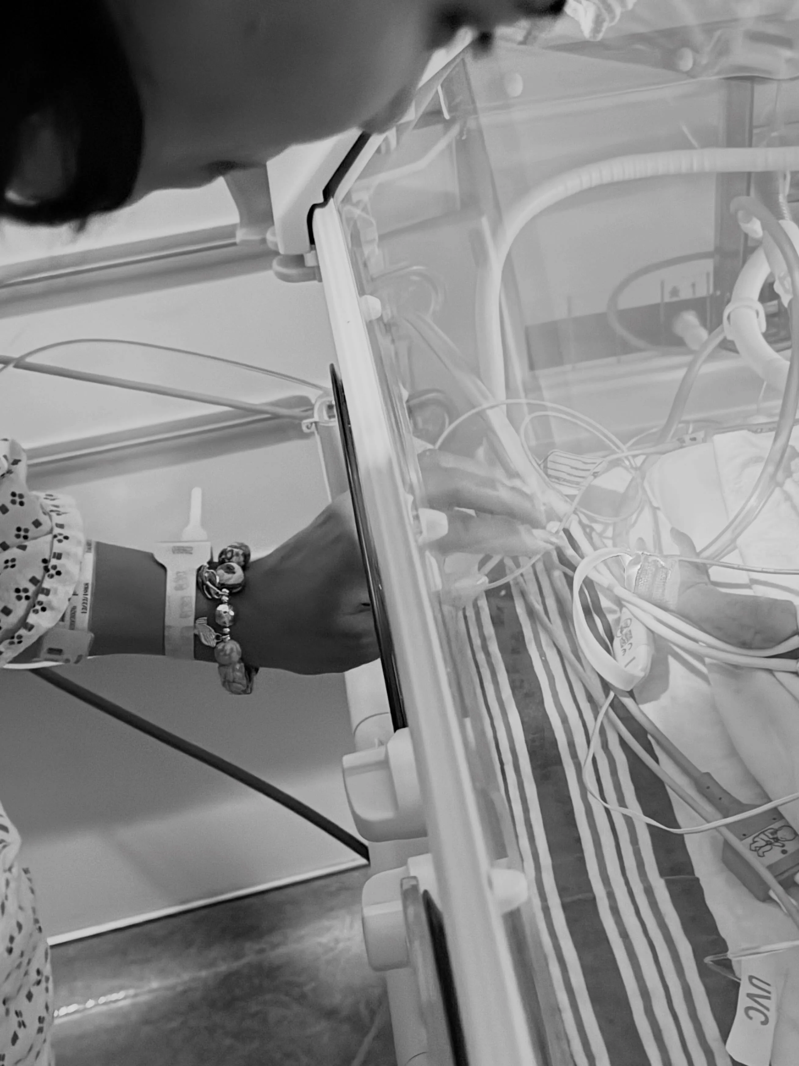 A person's hand with a hospital bracelet and beaded bracelet reaching into an incubator to touch a baby, with medical wires and striped bedding visible.
