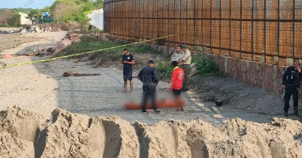 Law enforcement and emergency personnel gathered around a body on a sandy beach.