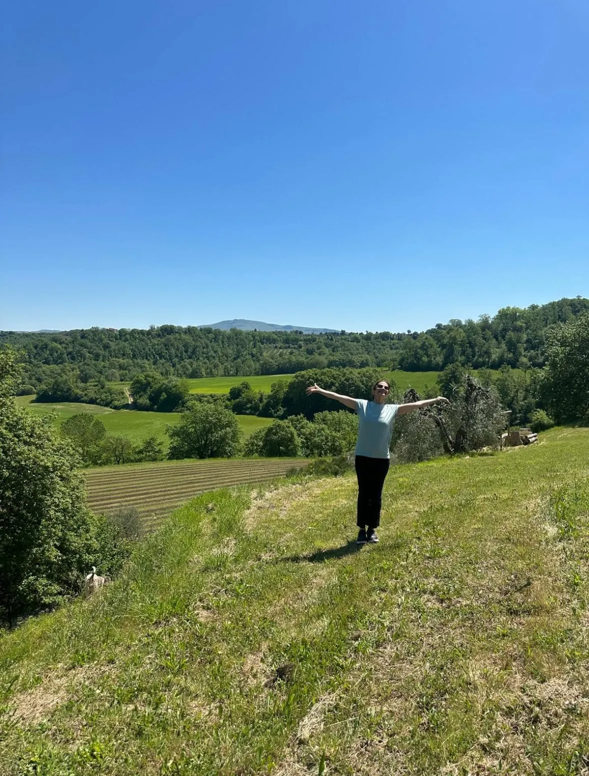 Livia Giuggioli Firth stands in a field with arms outstretched against a lush green valley with distant hills.