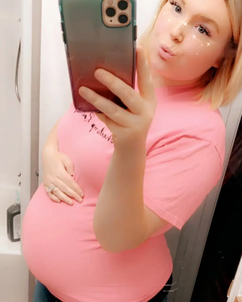 Reagan Hancock in a pink t-shirt showing her baby bump and making a kissy face while holding her phone.