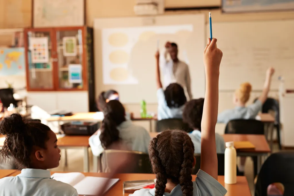 Rear view of a girl in a classroom raising her hand to answer a question.