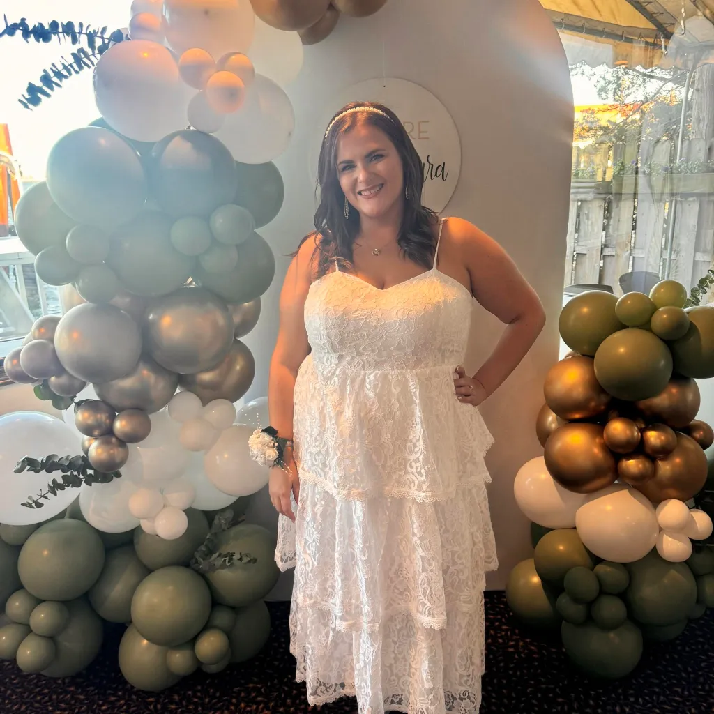 Woman in a white lace dress with a corsage, surrounded by green, white, and gold balloons.
