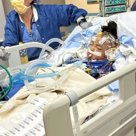 A young child lies in a hospital bed, hooked up to numerous medical devices, with a medical professional standing beside the bed.