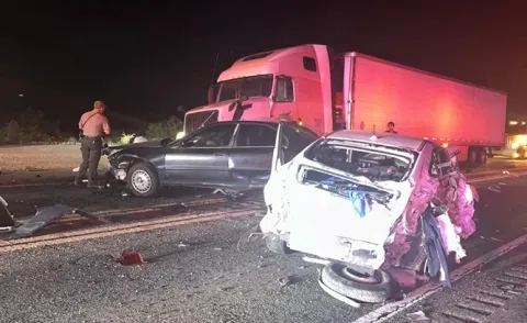 Nighttime image of a multi-car wreck involving a tractor-trailer and two severely damaged cars, with a person in a uniform standing nearby.