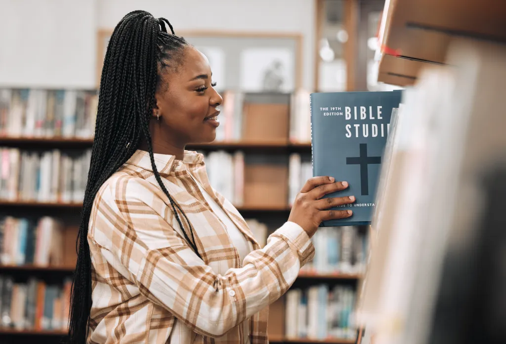 A smiling Black woman in a library examines a book titled 