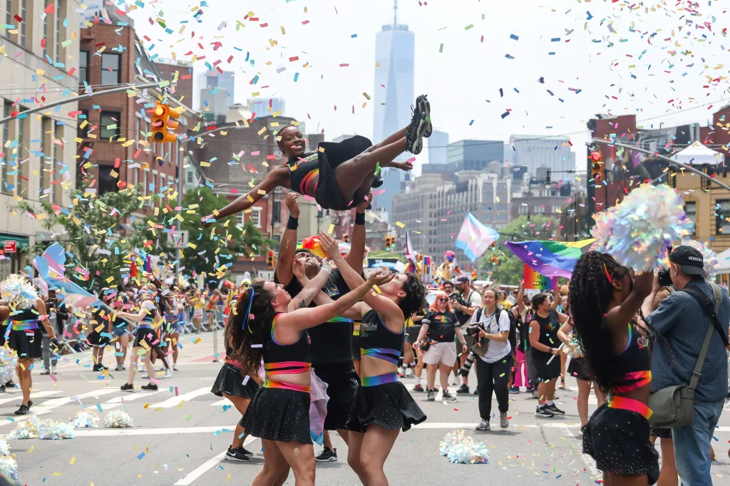A group of revelers in rainbow-colored attire toss a person into the air during the NYC Pride March, with confetti falling around them.