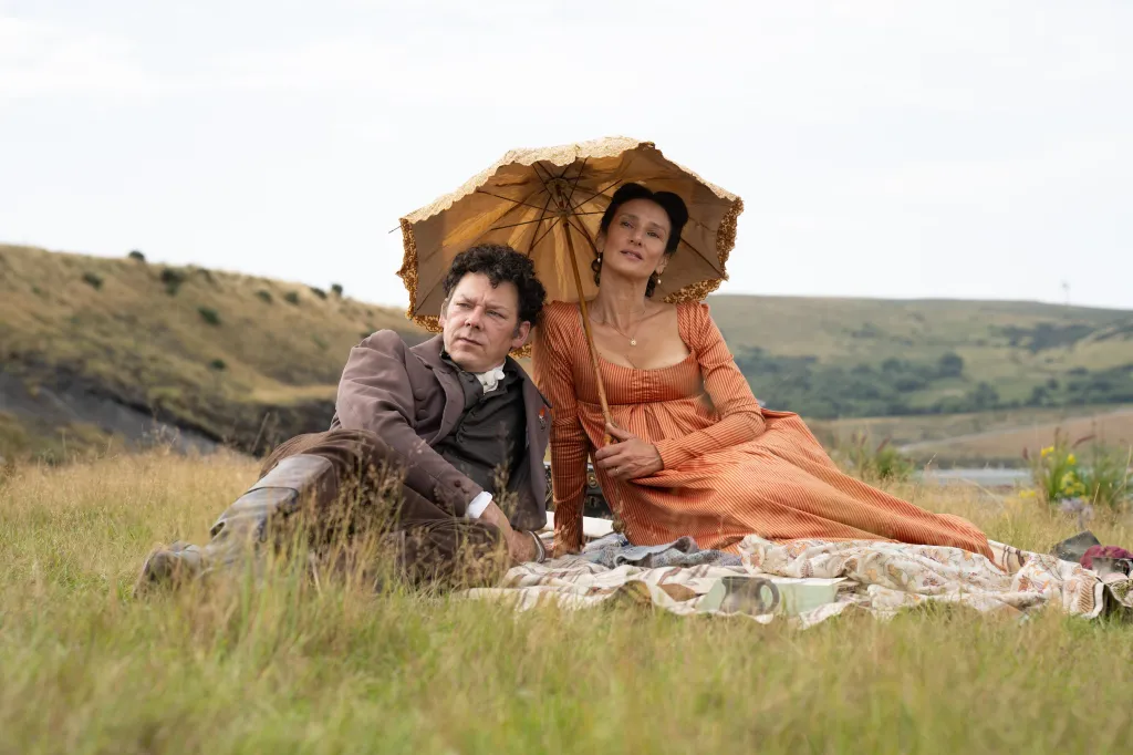 Richard Coyle and Indira Varma in period clothing, sitting on a blanket in a field under a parasol.