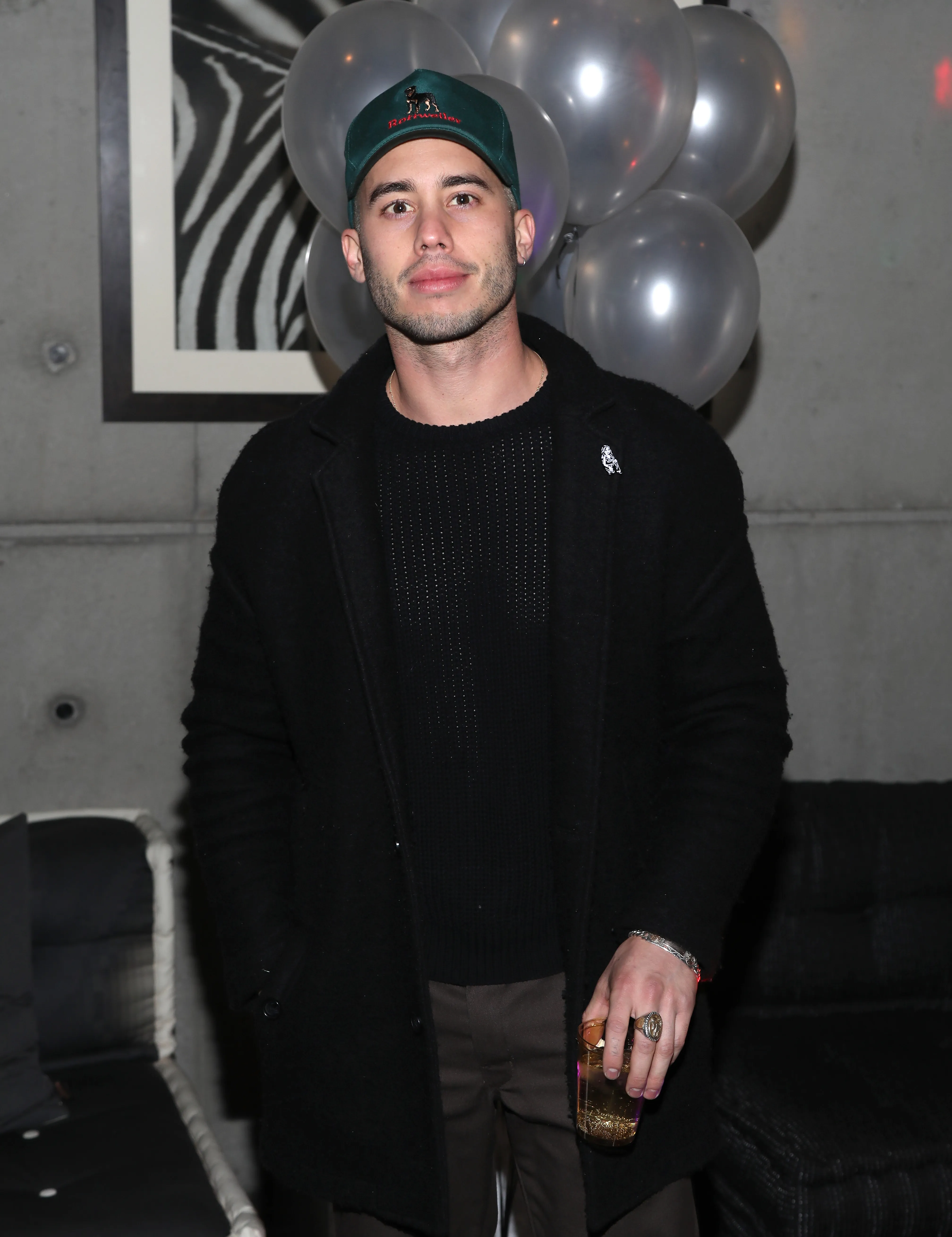 Ricky Alvarez in a black coat and green baseball cap, holding a drink, at a birthday party.