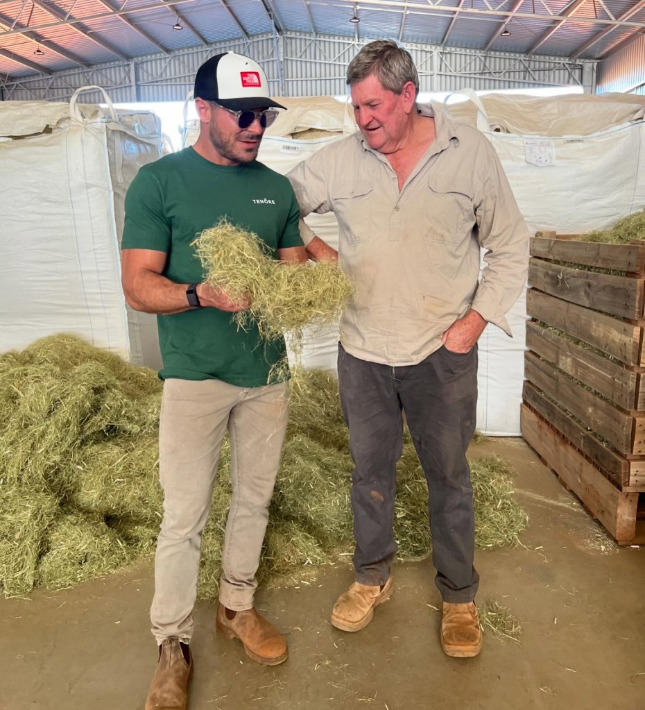 Zac Efron and Paul Rennie standing together, holding hay, on Rennie's farm in Hillston, NSW.