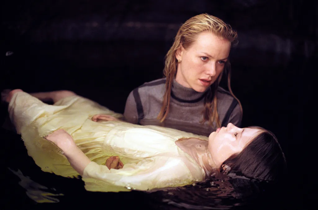 Naomi Watts with wet hair and a gray top looks at a young girl in a yellow dress floating in dark water in a scene from 
