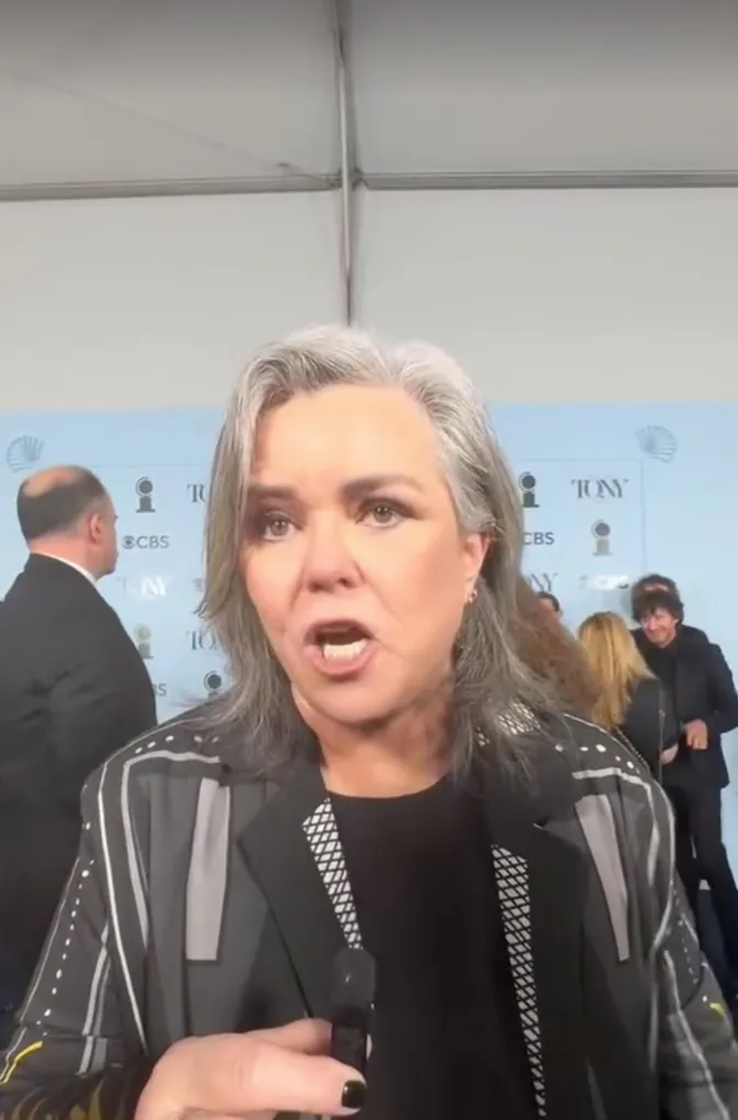 Rosie O'Donnell speaking to an off-screen person, with other people in the background.
