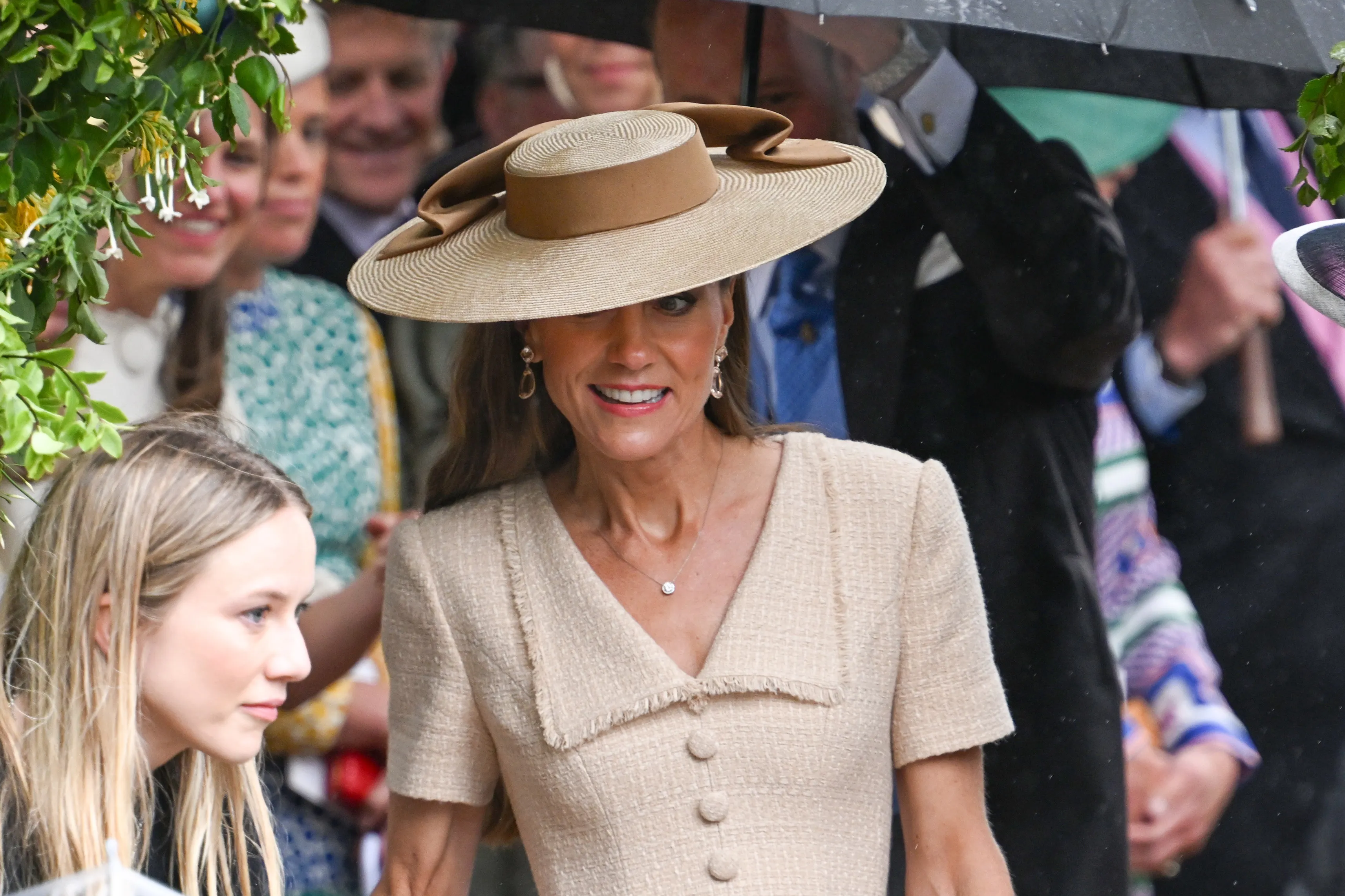 Catherine, Princess of Wales, smiles wearing a tan hat, matching dress, and diamond necklace.