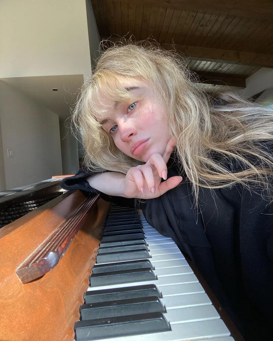 Sabrina Carpenter with blonde hair and bangs, resting her chin on her hand while leaning on a piano.