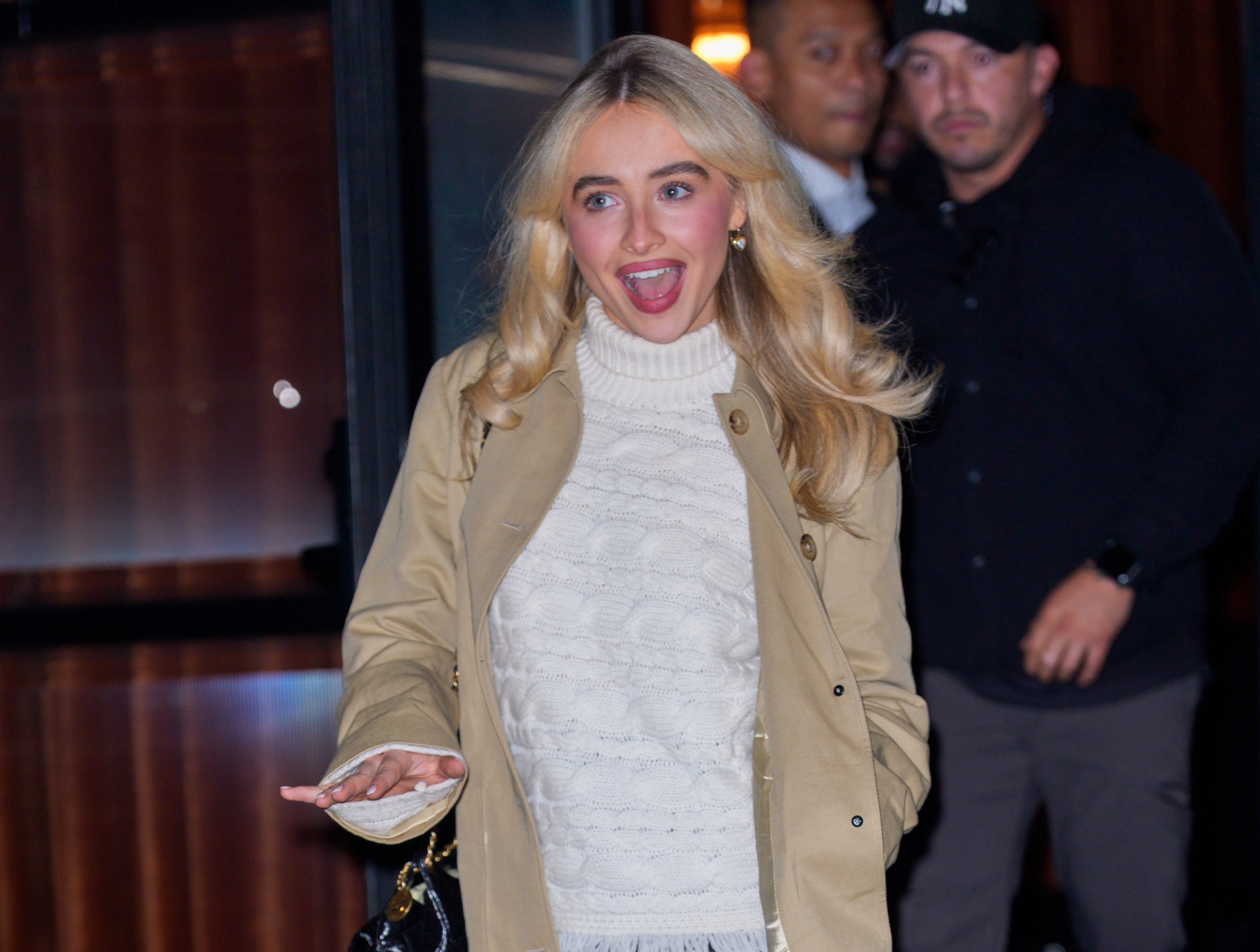 Sabrina Carpenter in a cream turtleneck and tan coat, looking surprised and animated.