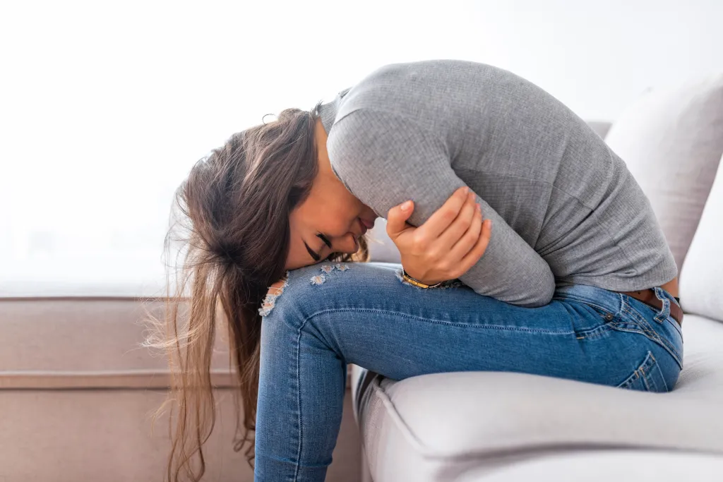 A woman sits on a couch, curled over her knees and holding her stomach, as if in pain.