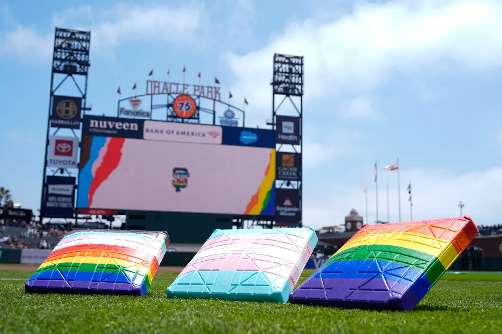 Three pride-themed bases on a baseball field at Oracle Park, with a jumbotron showing a San Francisco Giants logo with pride colors in the background.