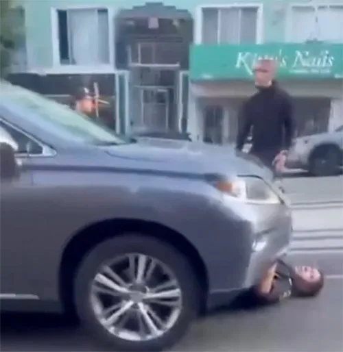 A person lying under the front of a gray car while another person stands behind the car.