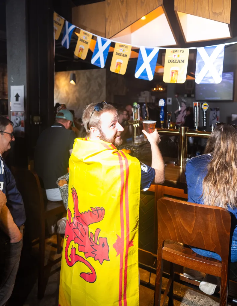 A man in a yellow and red Scottish flag, holding a drink, looks over his shoulder inside a bar decorated with Scottish flags.