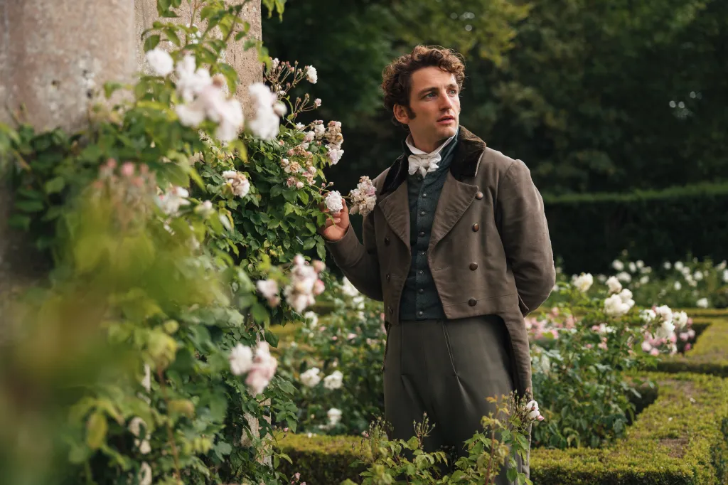 Laurie Davidson in period clothing, standing by a rose bush.