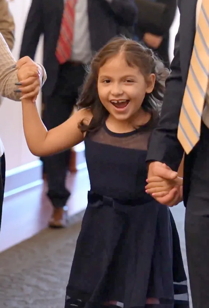 A smiling young girl in a dark dress holds hands with two adults.