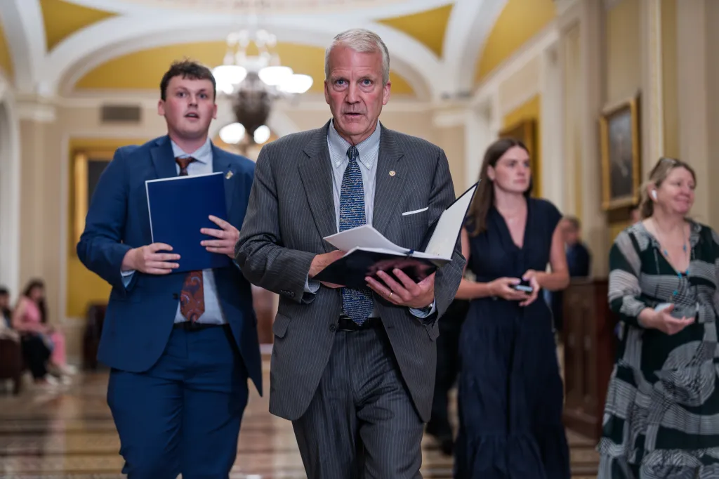 Sen. Dan Sullivan in a pinstripe suit and blue tie, walking with papers in hand, accompanied by other people in a hallway.