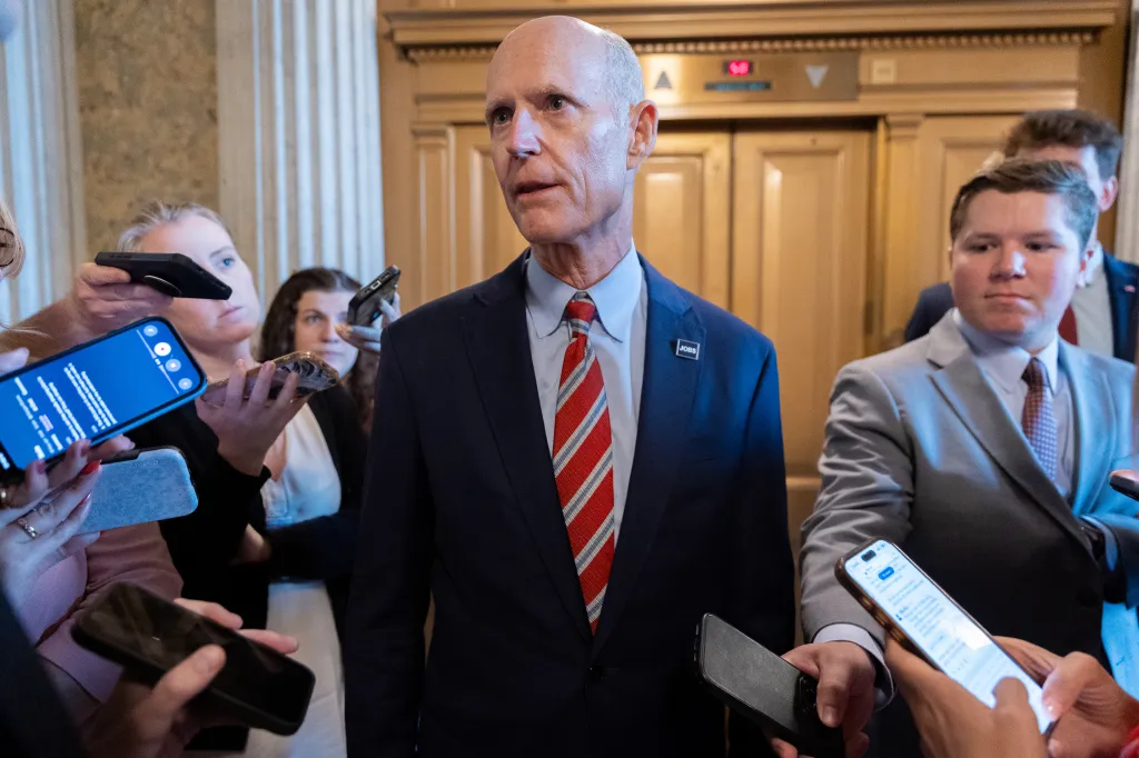 Senator Rick Scott speaks to reporters holding phones.