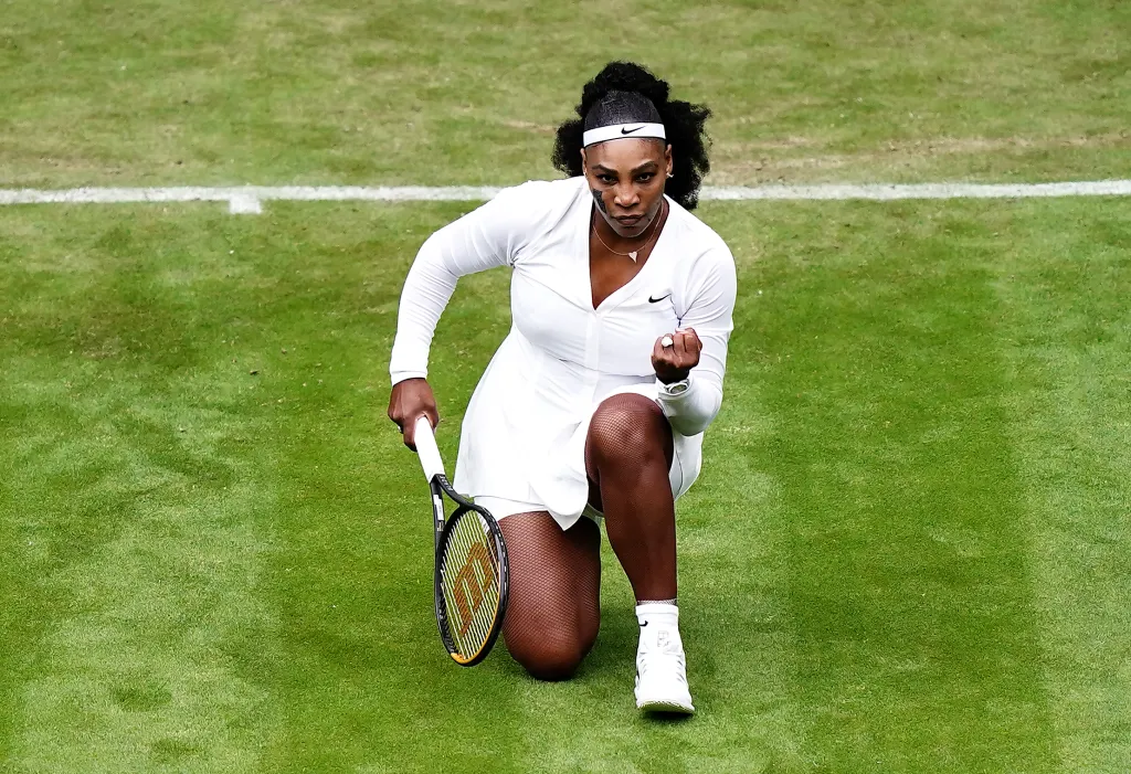 Serena Williams celebrates a break in serve, kneeling on a grass court with a clenched fist and holding a tennis racket.