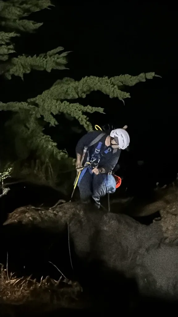 A firefighter in a helmet and harness rappels down a dark, rocky slope at night.