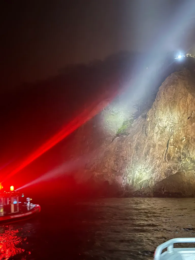 A rescue boat with red lights and spotlights illuminating a rocky cliff at night.