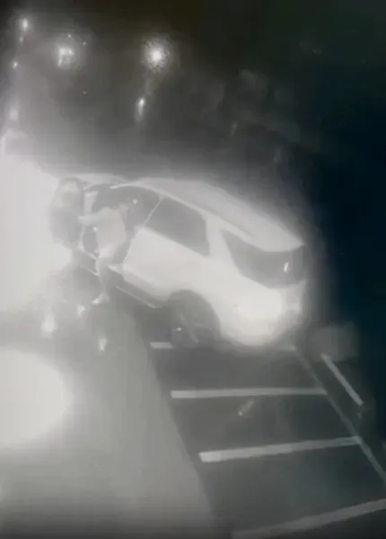 Grainy, dark footage of a white car with a door open, with a person appearing to exit the vehicle and another figure on the ground.