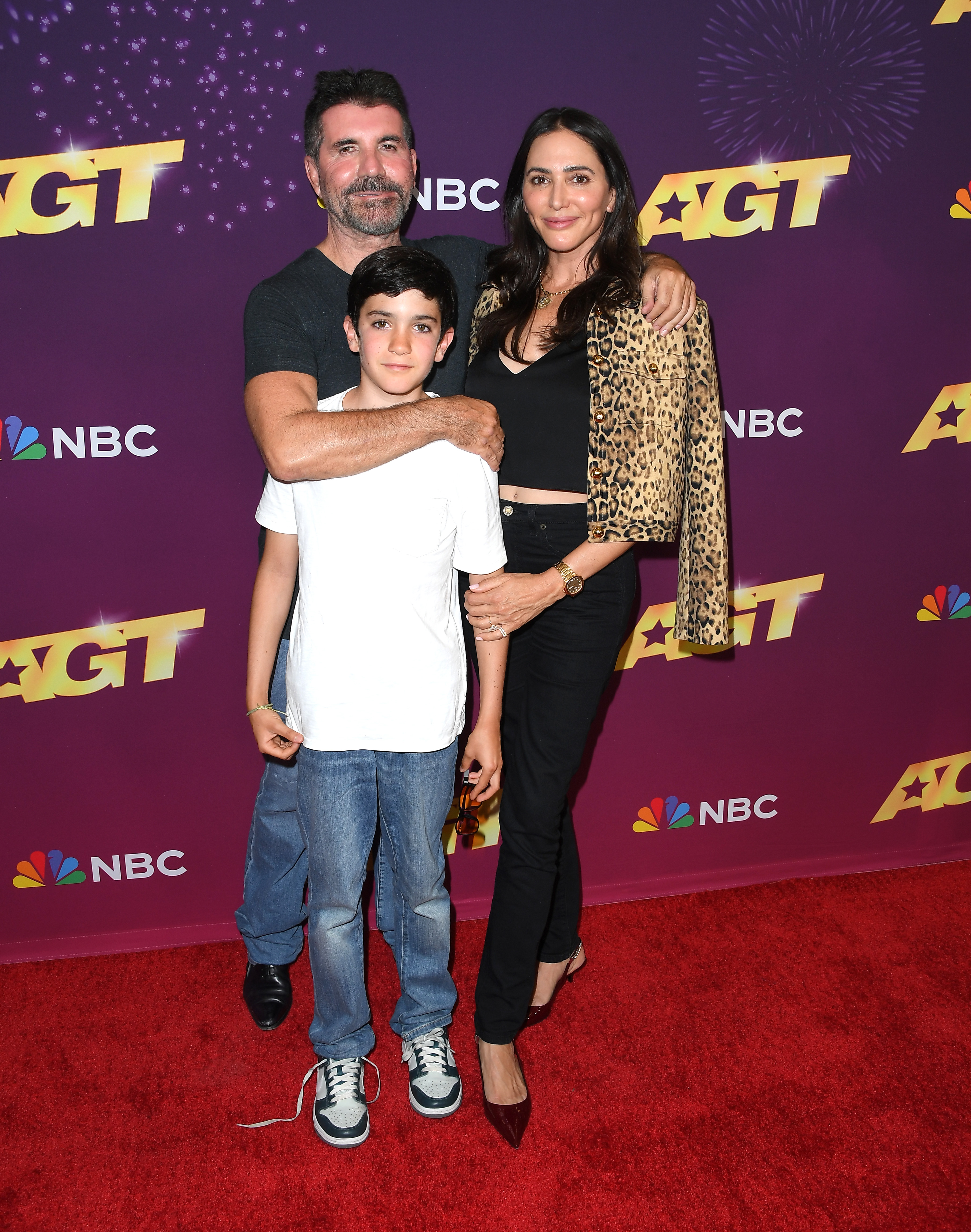 Simon Cowell, his son Eric Cowell, and Lauren Silverman pose on a red carpet in front of a purple backdrop with 