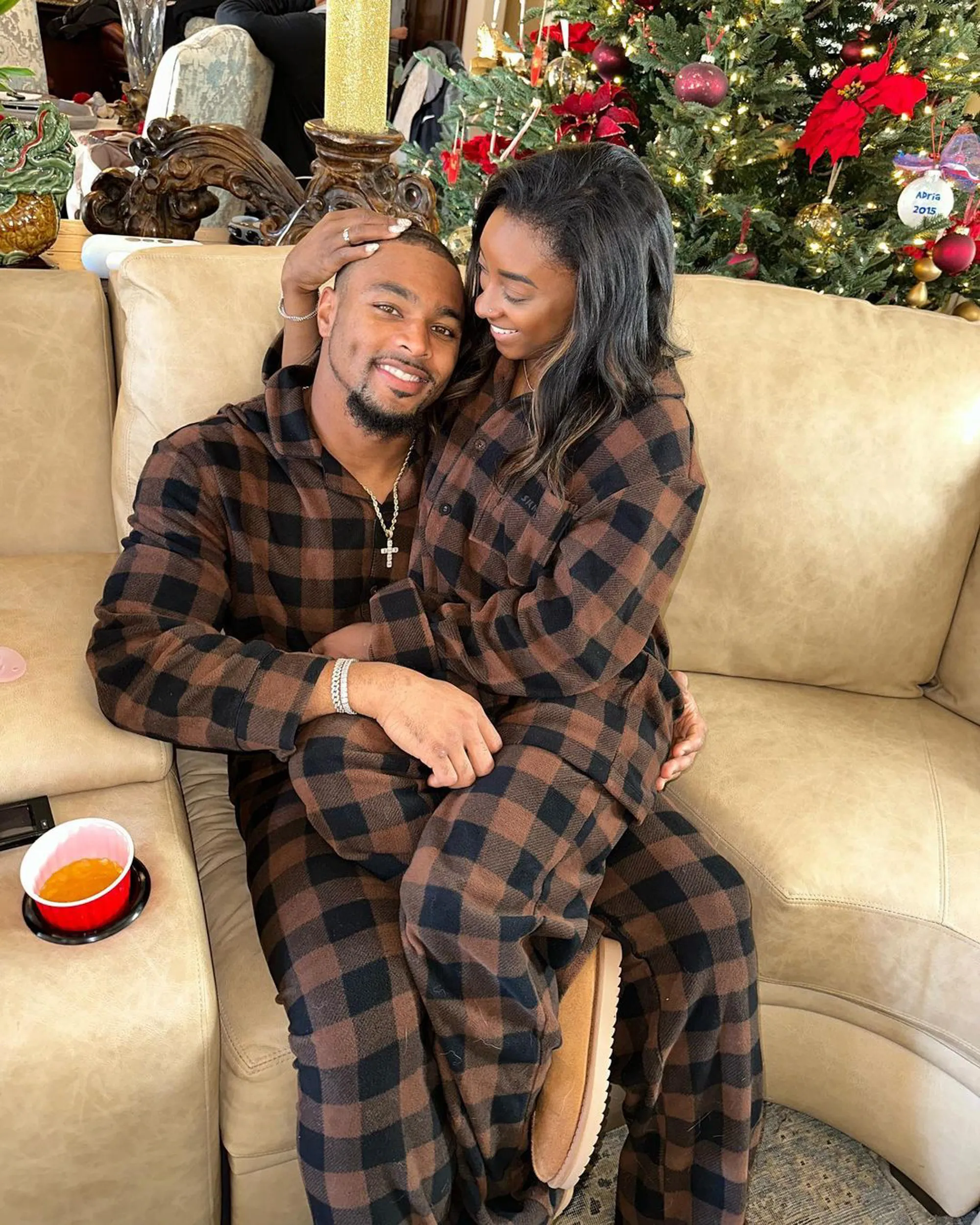 Simone Biles and Jonathan Owens sitting on a couch in matching brown and black plaid pajamas.